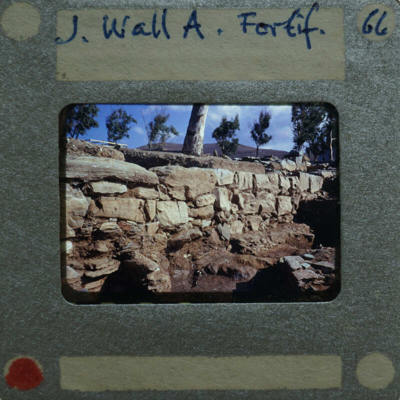 Old photograph of a stone wall with trees and sky in the background, on a film strip with handwritten details.