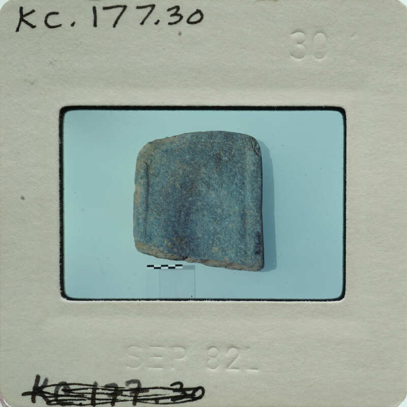A small, dark blue rock on a white surface with a label and date stamp.