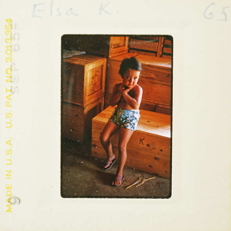 A young child standing with hands on hips, wearing shorts and flip-flops, next to a wooden bench.