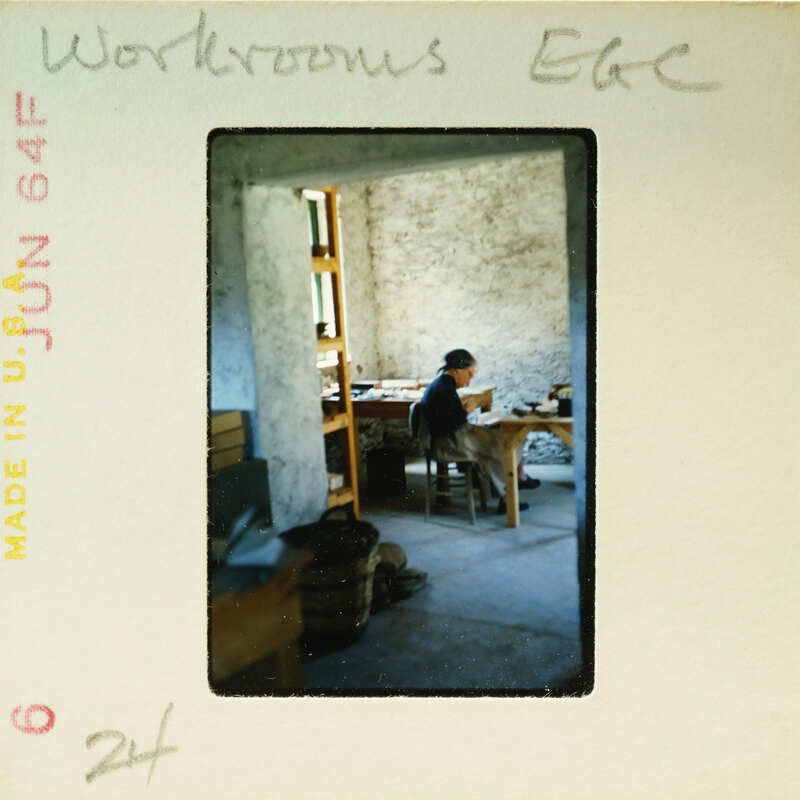 A person sitting at a desk with papers and a book, surrounded by shelves and boxes in an old room.