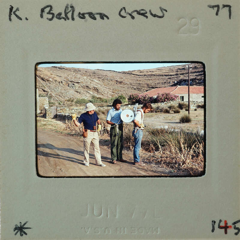Three men standing on a dirt road with mountains and a house in the background.