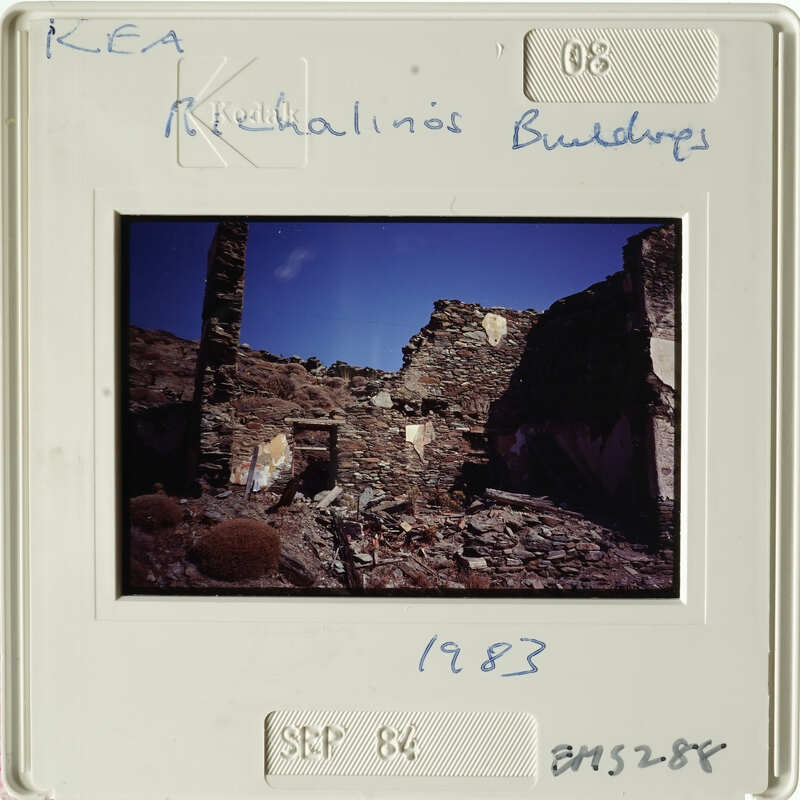 An old photograph of a ruined building with blue sky and bushes in the foreground, dated 1983.