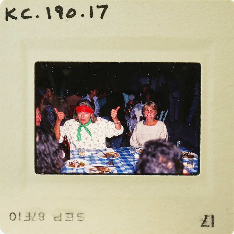 Two women sitting at a table with food, smiling and giving thumbs up. Catalog number KC.190.17