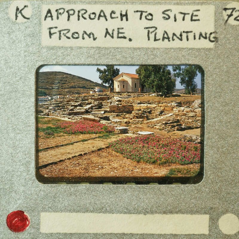 A photograph of an archaeological site with ruins and a small building, taken from the approach to the north-eastern entrance.
