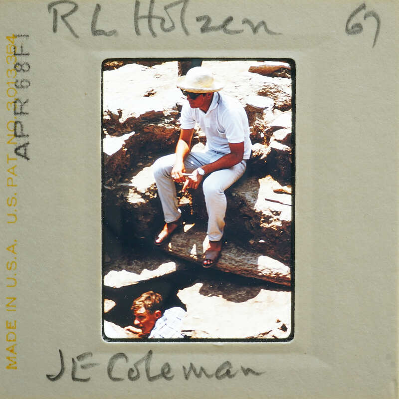 Man sitting on rock with another person behind him, wearing a hat and light-colored clothing.