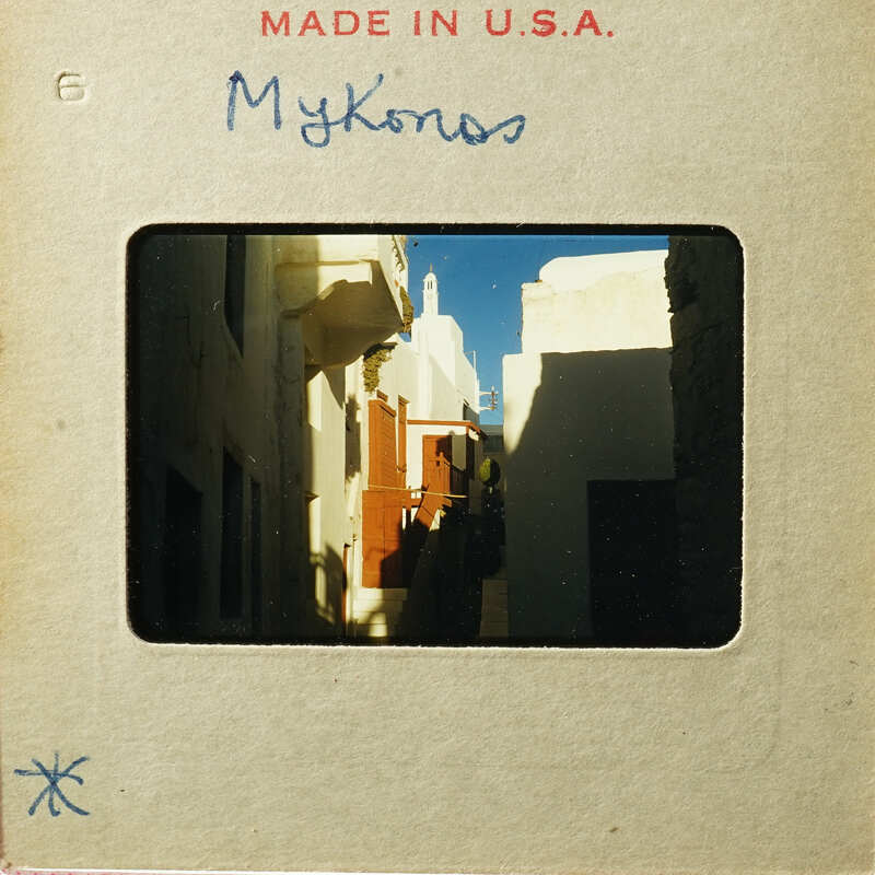 Old photograph of a narrow street with buildings and blue sky, labeled "Mykonos" on a cardboard mount.