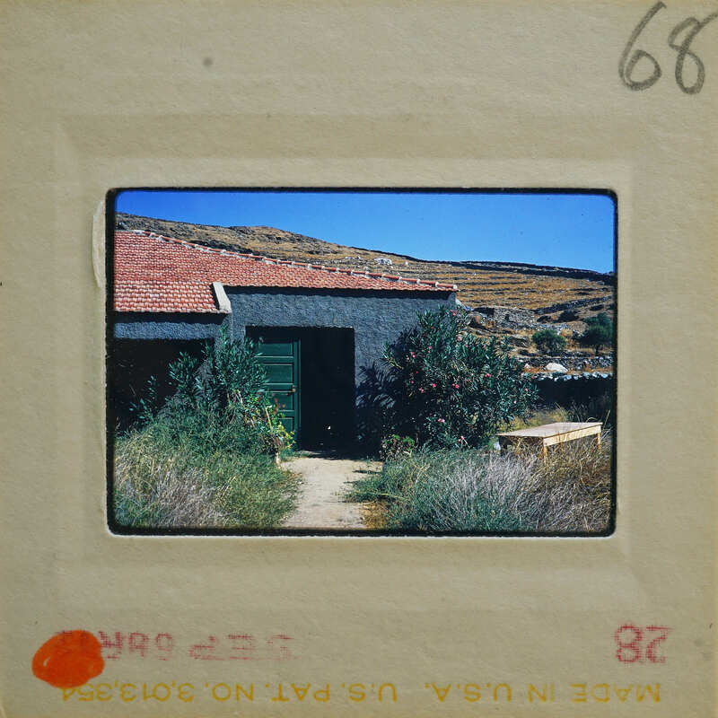 A photograph of a rustic house with a red tiled roof, surrounded by greenery and mountains in the background.