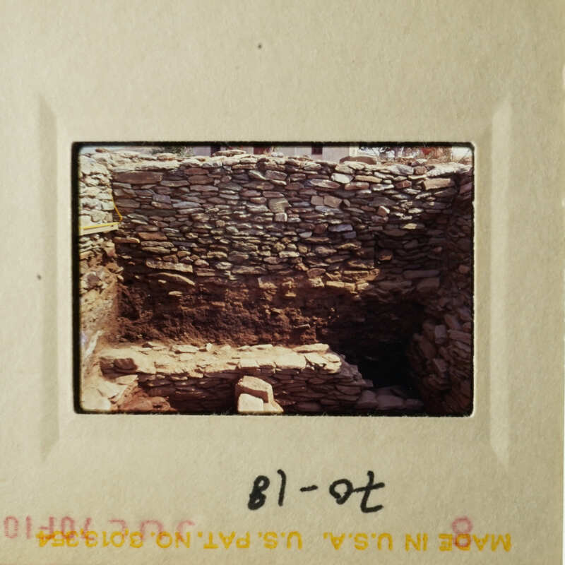 The photo shows an ancient stone structure, possibly ruins, with a rock wall and a dirt floor.