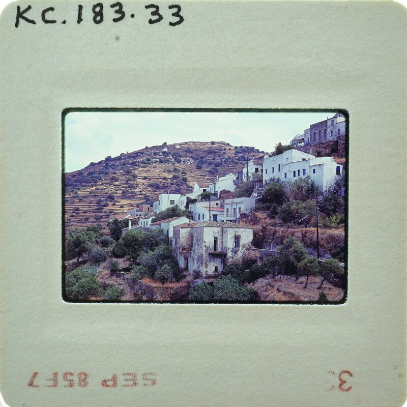 A vintage photograph of a mountainous village with white houses and green vegetation, taken from a distance.
