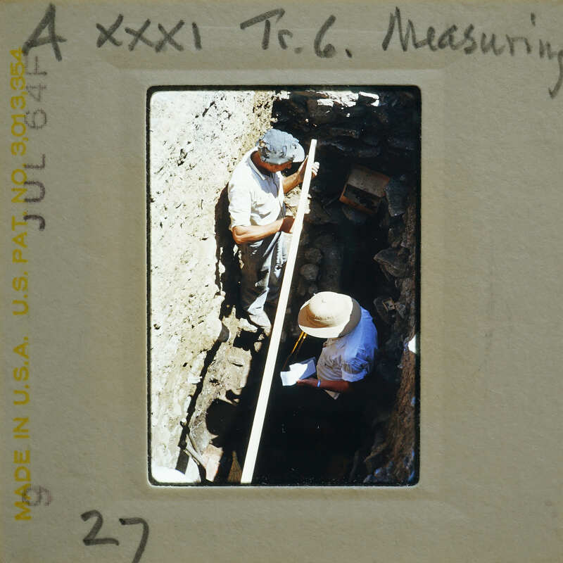 Two people working on a archaeological site with excavated earth and ruins visible.