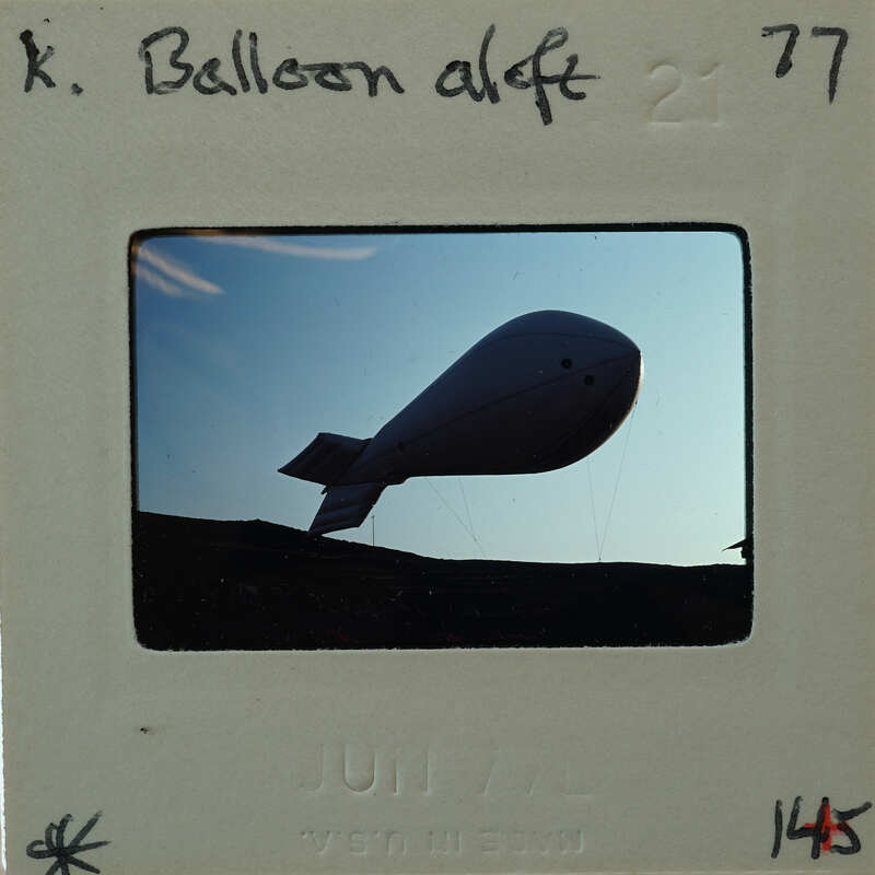 A black and white photograph of a large, round balloon with a tail flying against the sky.