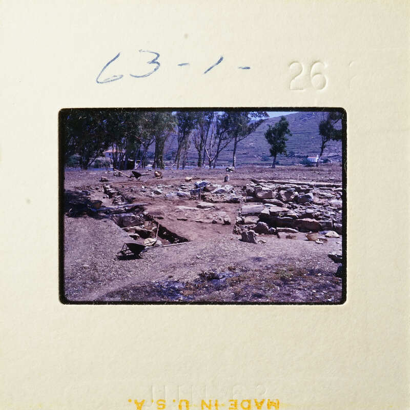 Ancient ruins with stone walls and foundations, set against a backdrop of trees and hills.