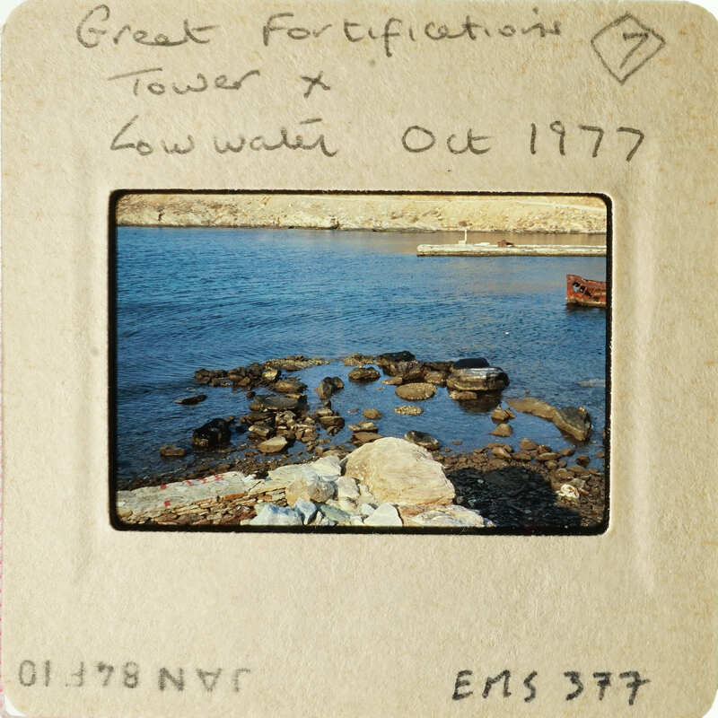 A photograph of an old fortification by water, possibly a harbor, with low tide exposing rocks and seaweed.