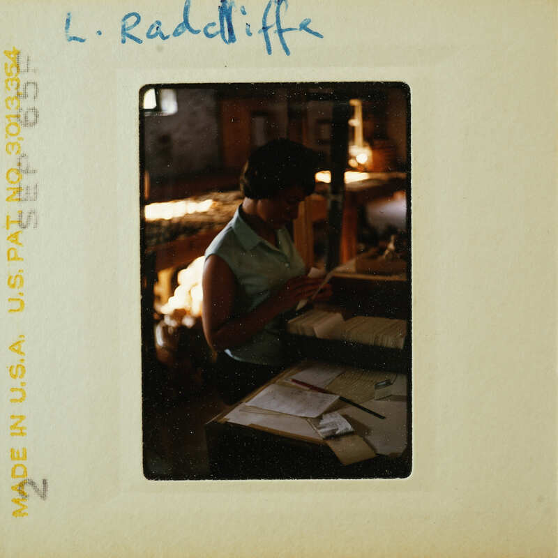 A vintage photograph of a woman sitting at a desk with papers and a pen, wearing a light-colored blouse.