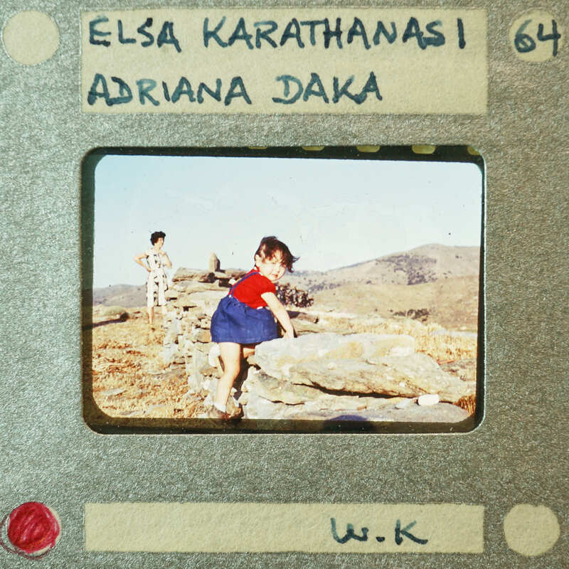 A vintage photo of a child standing on rocks with mountains in the background, labeled "Elsa Karathanasi" and "Adriani Daka."
