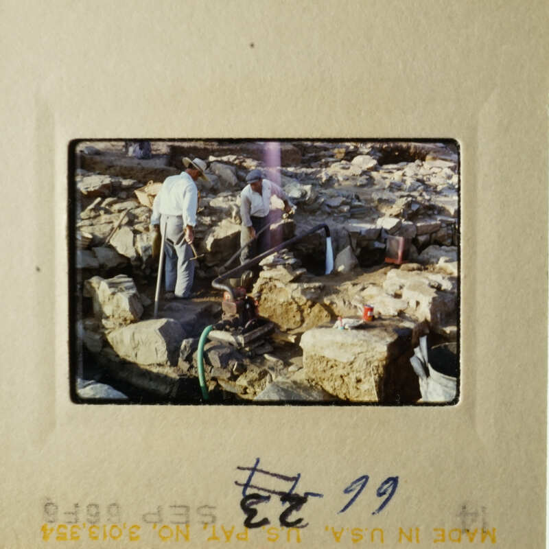 Image depicts an outdoor scene with two men working on a construction site, surrounded by large rocks and tools.