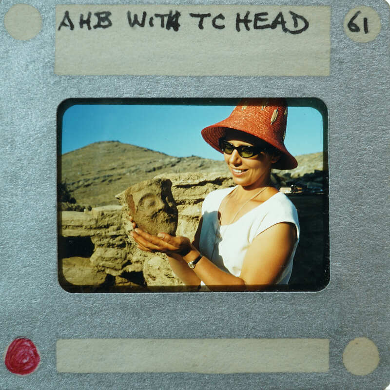 A woman holding a terracotta head (K1.458) with mountains in the background.