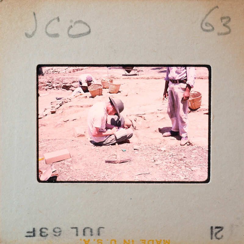 A man sitting on the ground, looking at something small in his hands while another person stands nearby.