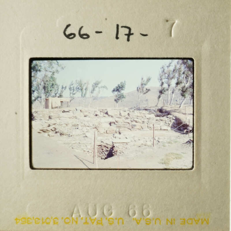 This photo shows an old, faded photograph of a barren landscape with a dirt road and some small structures, possibly ruins, under a clear sky.