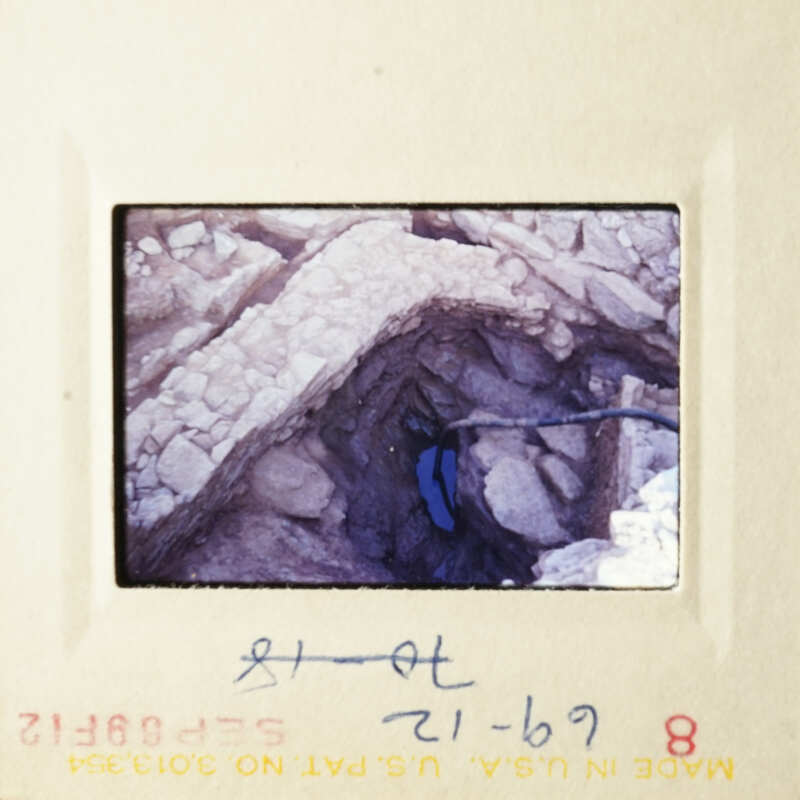 This is a photograph of an old photo, showing a rocky cave with a small opening and a handwritten note.