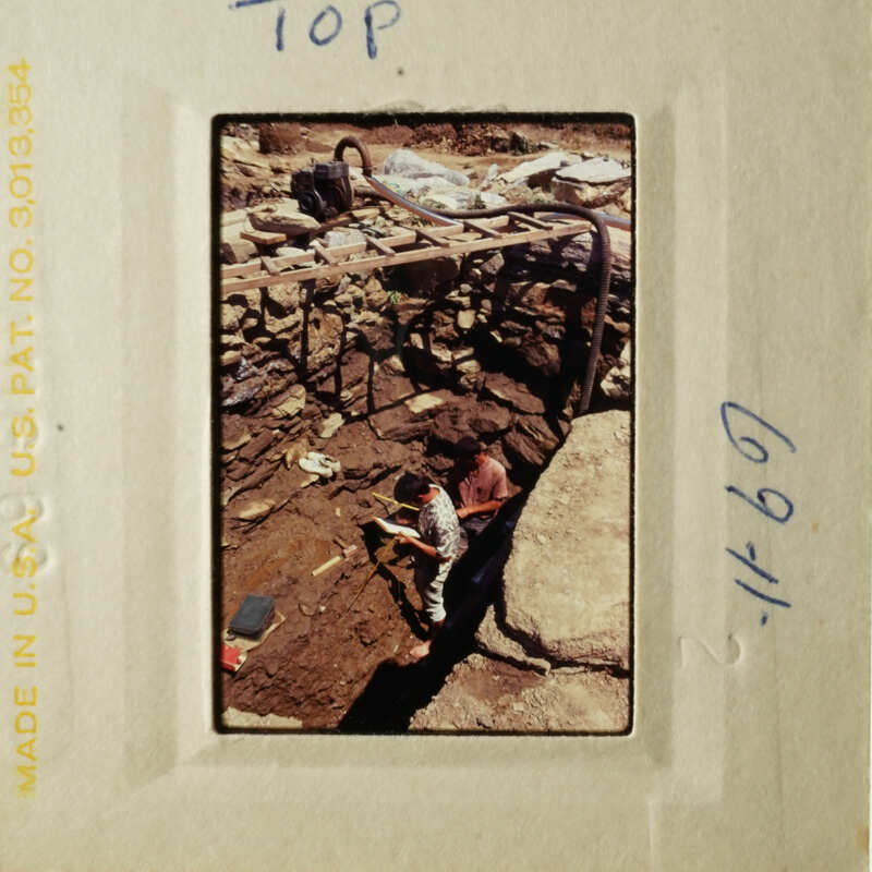 A vintage photo showing a person working on an excavation site with a shovel, surrounded by construction equipment and materials.
