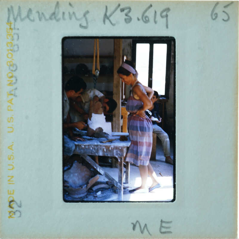 Woman standing at a workbench with two men, who are restoring an artifact.