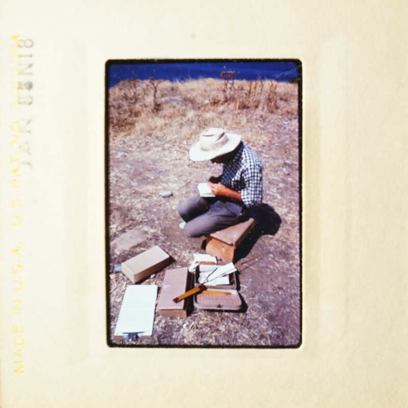 Man sitting on ground with papers and tools, outdoors.