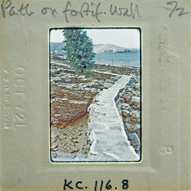A vintage photo of a stone path leading down to the water, with mountains and trees in the background.