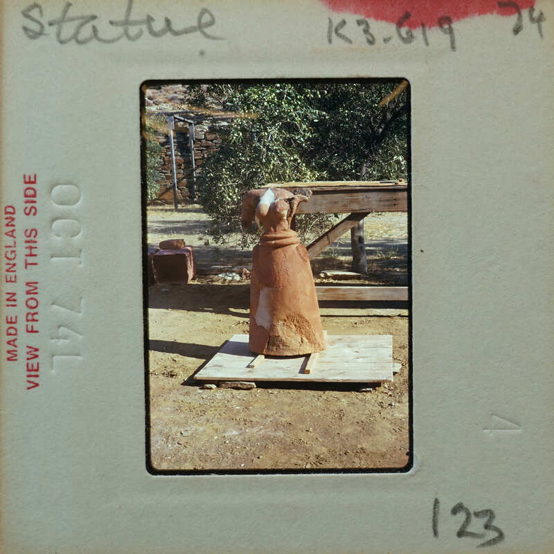 Terracotta figurine body sitting on wooden platform with trees and building in background, K3.619.