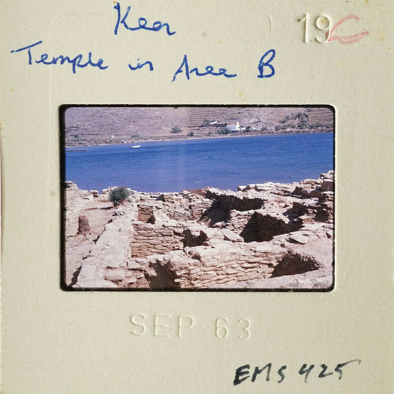 A photograph of an ancient temple ruins by a body of water with blue sky and mountains in the background.
