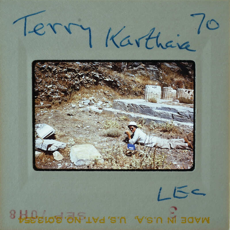 A vintage photo of a person sitting on the ground with an ancient stone structure behind them, labeled "Terry Karthaia 70" and marked with film strip details.