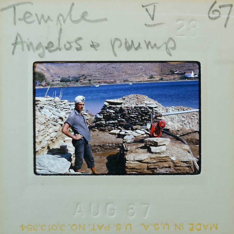 A man standing next to a stone wall and water, with a water pump on one wall and a bay in the background.