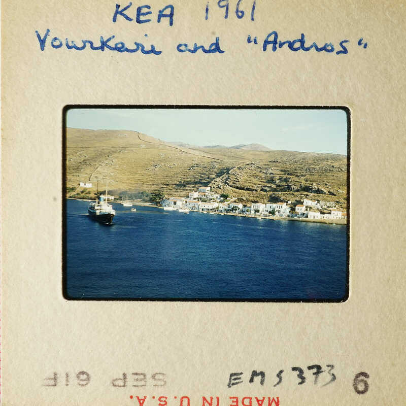 A vintage photograph depicting a coastal town with boats on the water, set against a backdrop of mountains and clear skies.
