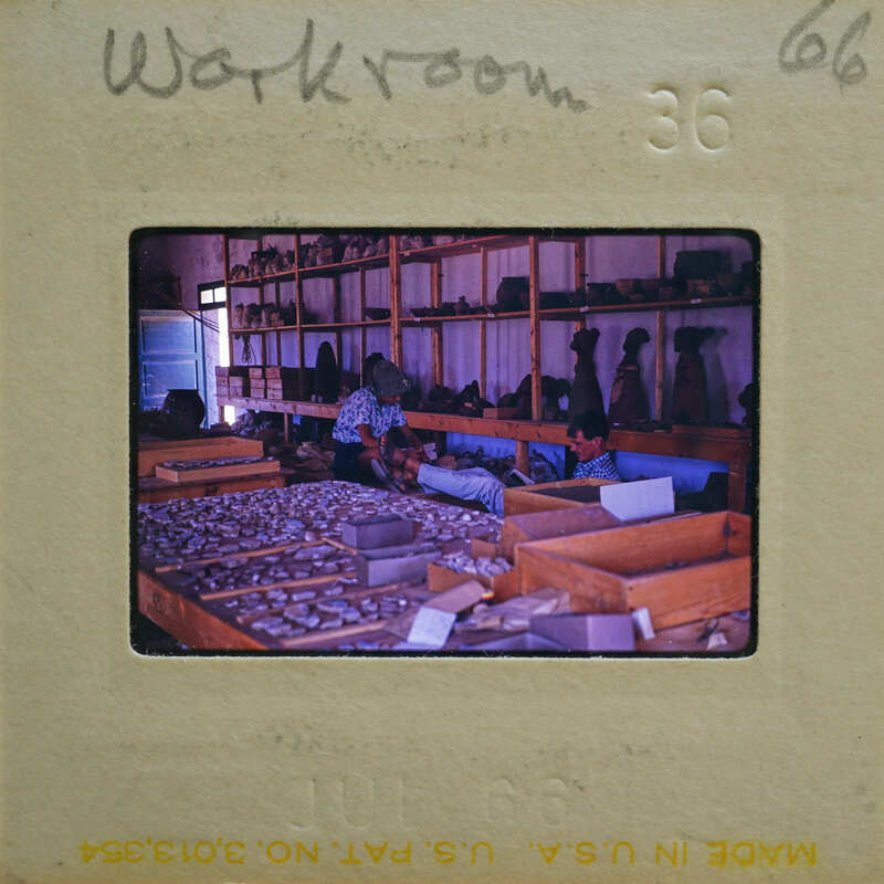 This image depicts a workshop with wooden shelves and various items, where two individuals are seated at workstations.