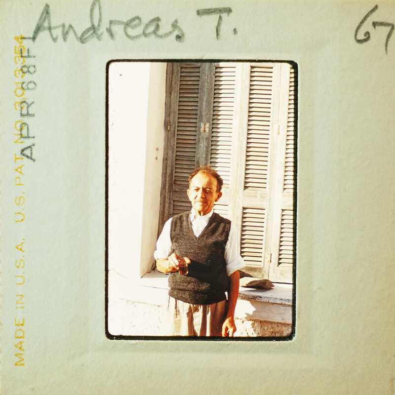 Old man wearing a sweater vest and standing outside with shuttered windows behind him.