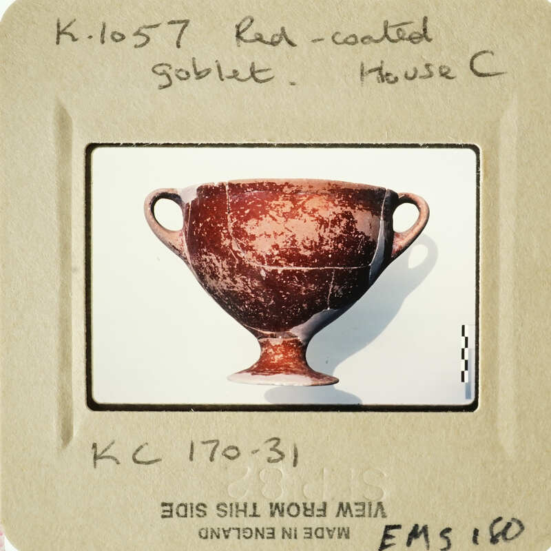 Ancient Greek-style cup with red coating and two handles, displayed against white background.