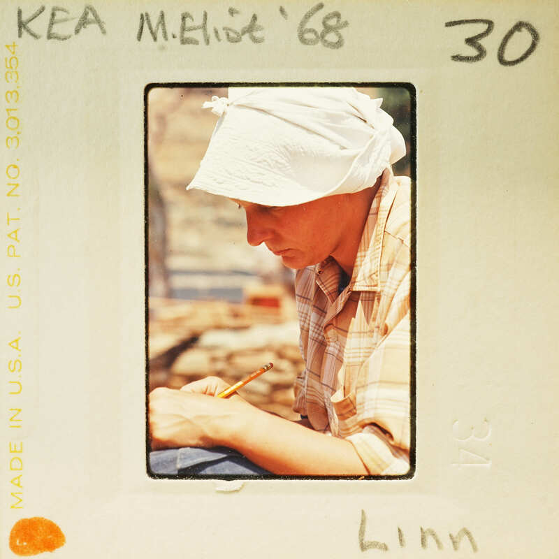 Woman sitting down writing with pencil and paper, wearing a hat.