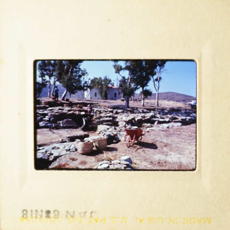 This is an image of a vintage photograph depicting a rural scene with a stone house, trees, and a person sitting on a chair.