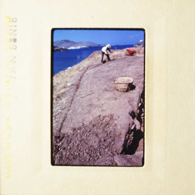 A person working on a dirt path near the sea, with a basket and a shovel nearby.