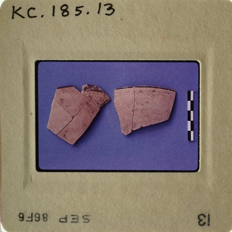 Two broken pottery shards on blue background with white border, labeled "KC.1856.13".