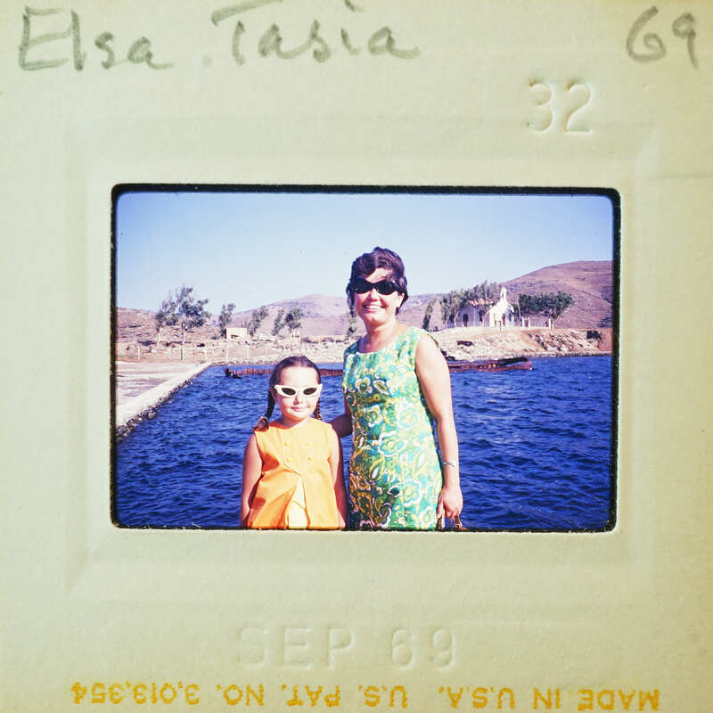 Two women posing together on a pier with water and mountains in the background.