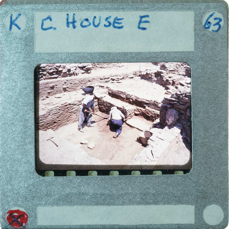 A photograph of two people working at an archaeological site with walls and a doorway.