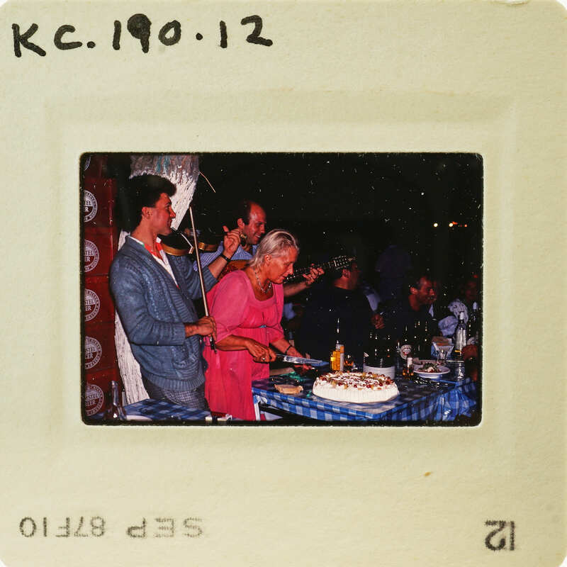 A vintage photograph of a group of people gathered around a dining table with a cake, celebrating an event. Catalog number KC.190.12