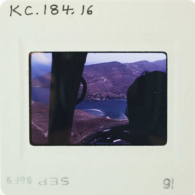 A vintage photograph of a person sitting inside a helicopter, looking out over a scenic landscape with mountains and water.