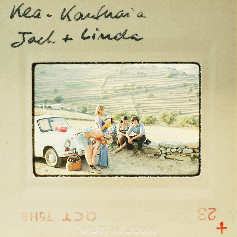 A vintage photo of a group of people sitting on a wall with a car and mountains in the background.