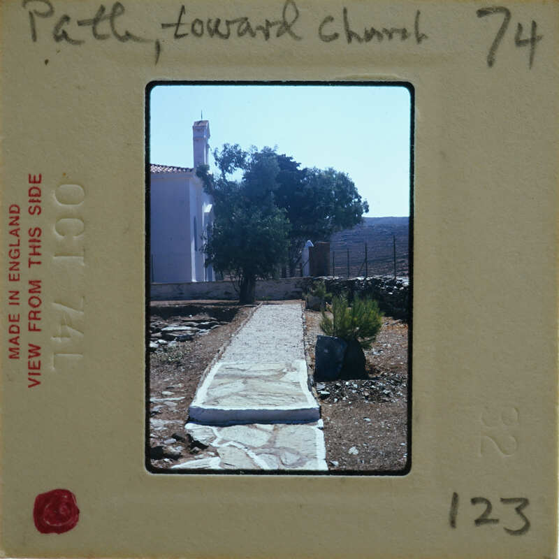 The image shows a vintage photograph of a stone path leading towards a building with trees and shrubs, set against a clear sky.