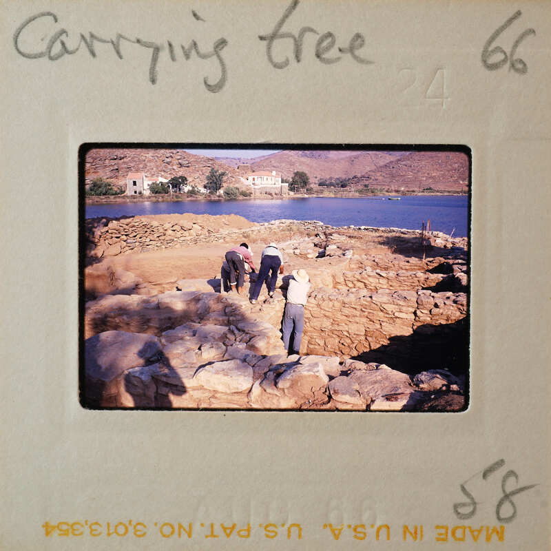 A photograph of people moving a tree trunk near water, with mountains in the background.