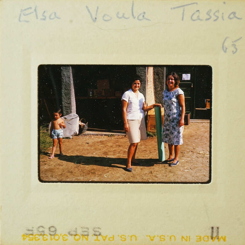 Two women standing together, smiling at the camera with a young child and an adult male behind them.