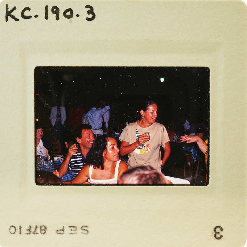 A vintage photograph of a group of people gathered at an event, with some individuals standing and others seated around tables. Catalog number KC.190.3