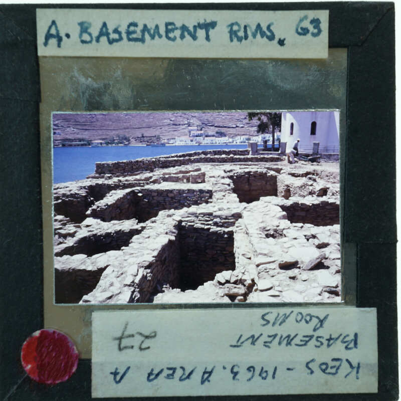 A photograph of a historical stone structure, possibly ruins, with the sea and mountains in the background.